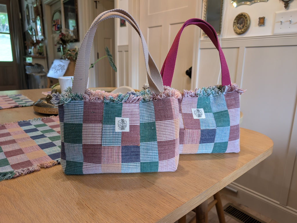 Two handwoven checkered tote bags in soft pink, teal, and blue tones with fringe trim, displayed on a wooden table