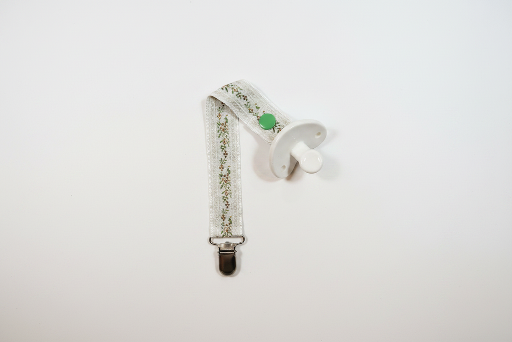 Handmade cream botanical pacifier clip with green snap button and silver metal clip on white studio background