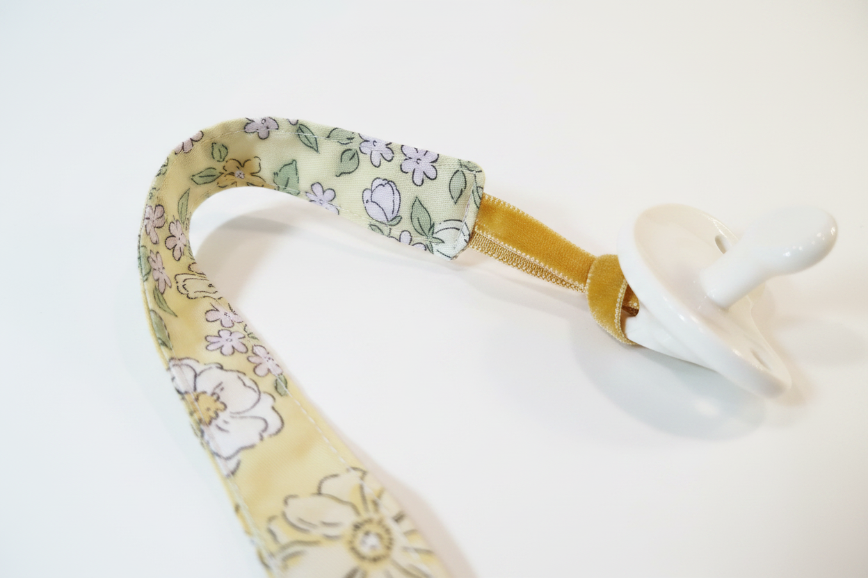 Close-up detail of handmade golden floral pacifier clip showing reversible design, brightened on white studio background