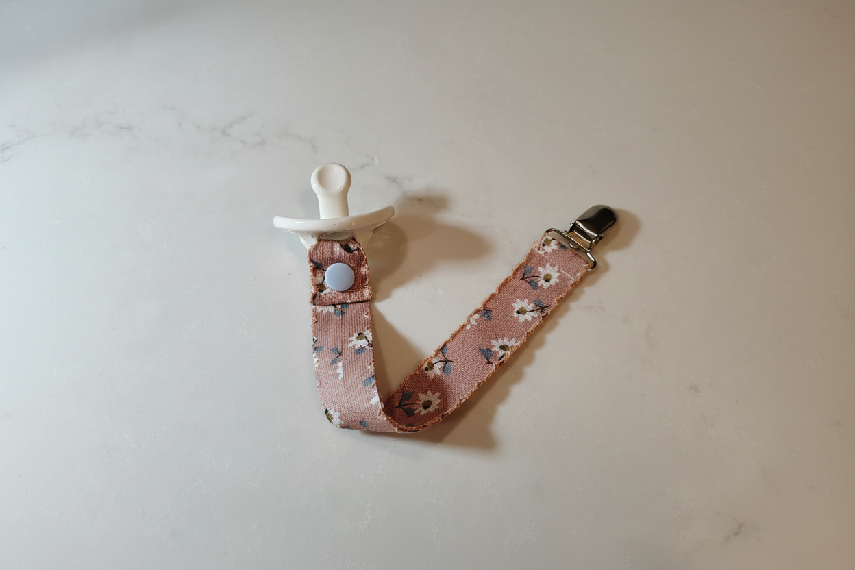 Handmade dusty rose daisy pacifier clip with blue snap button and silver metal clip on white studio background