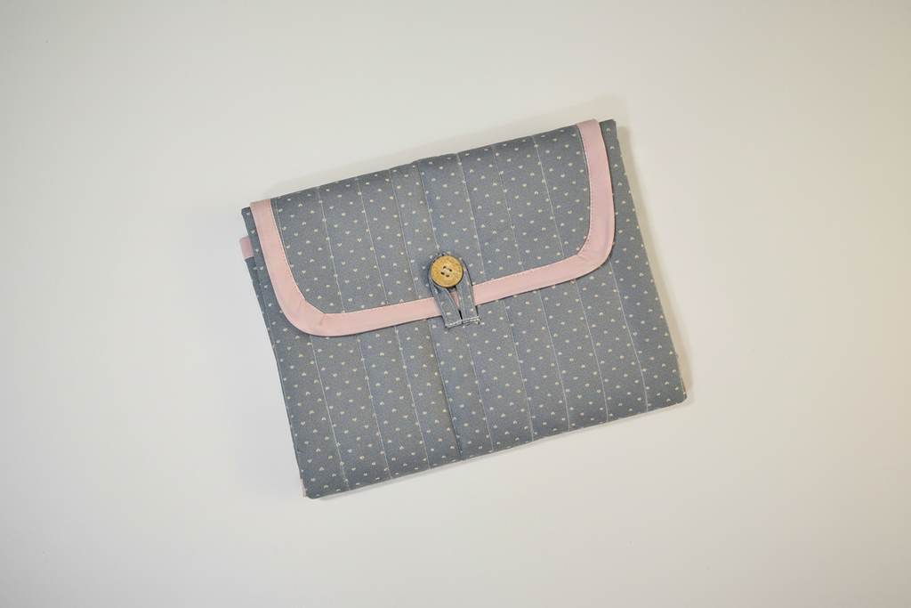 Handmade quilted changing mat in dusty blue-grey with white floral print, pink trim and gold button closure on white studio background