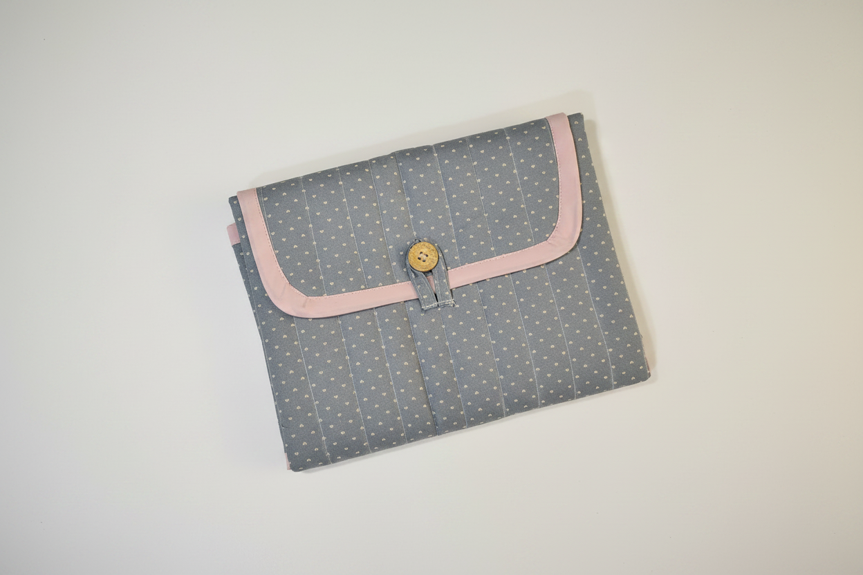 Handmade quilted changing mat in dusty blue-grey with white floral print, pink trim and gold button closure on white studio background
