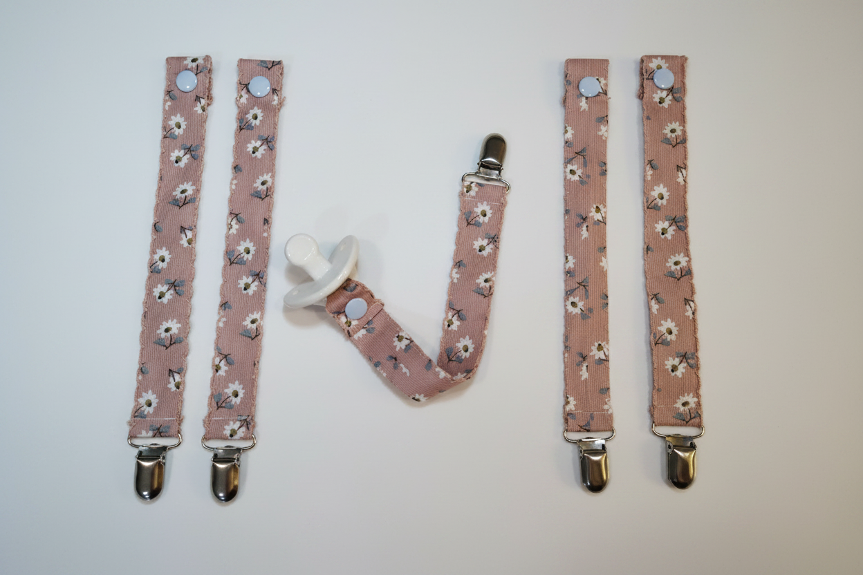 Collection of handmade dusty rose daisy pacifier clips and straps with silver metal clips on white studio background